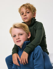 Two young boys pose together against a plain background. One wears a dark green Jacket – Merino Kids AirWool zip-up top and rests his arm on the other, who wears a blue Jacket – Merino Kids AirWool zip-up top and sits in front. Both look into the camera with neutral expressions.