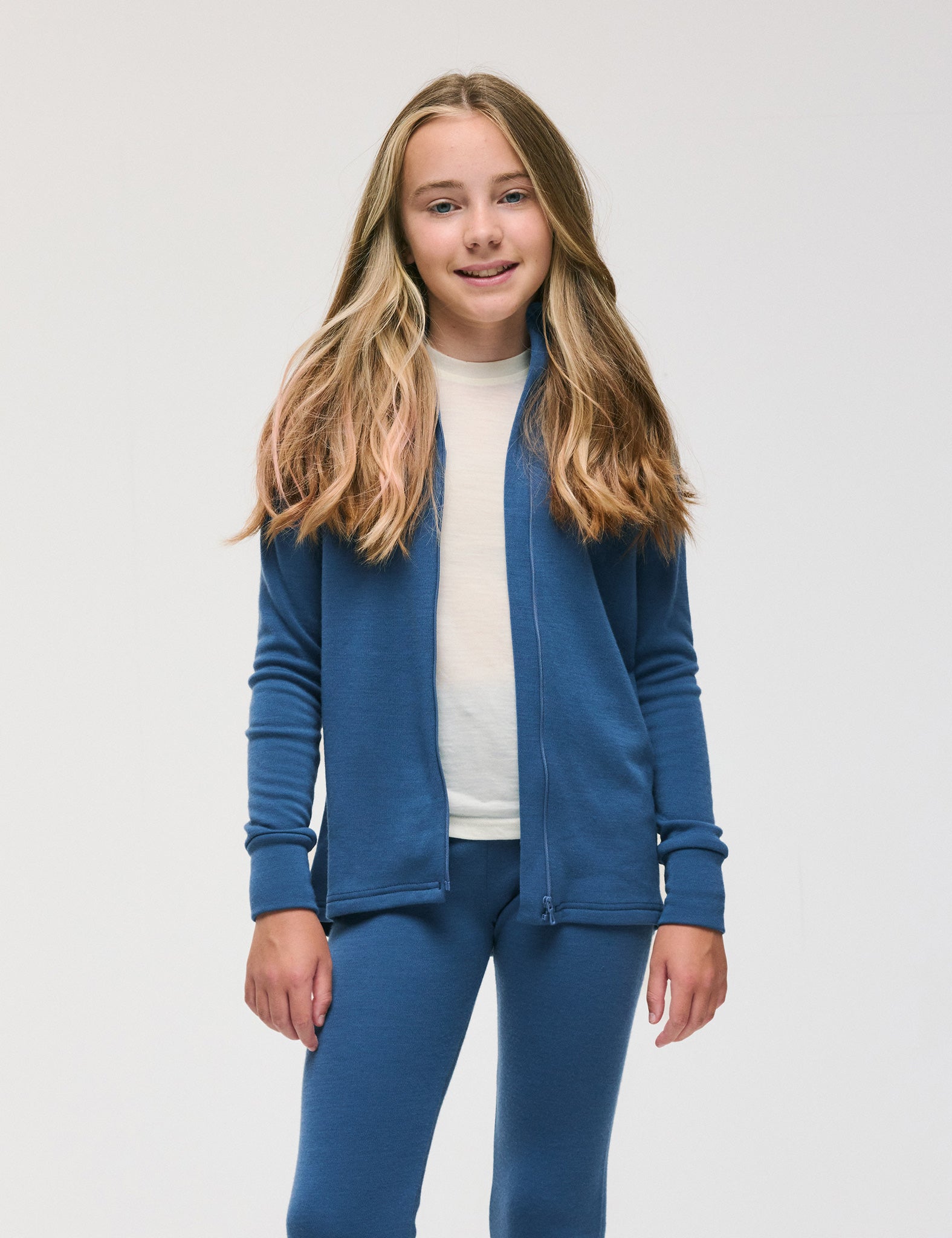 A young girl with long, light brown hair stands and smiles against a plain white background, wearing a blue Merino Kids AirWool jacket and matching blue pants over a white shirt.