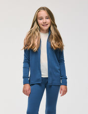A young girl with long, light brown hair stands and smiles against a plain white background, wearing a blue Merino Kids AirWool jacket and matching blue pants over a white shirt.