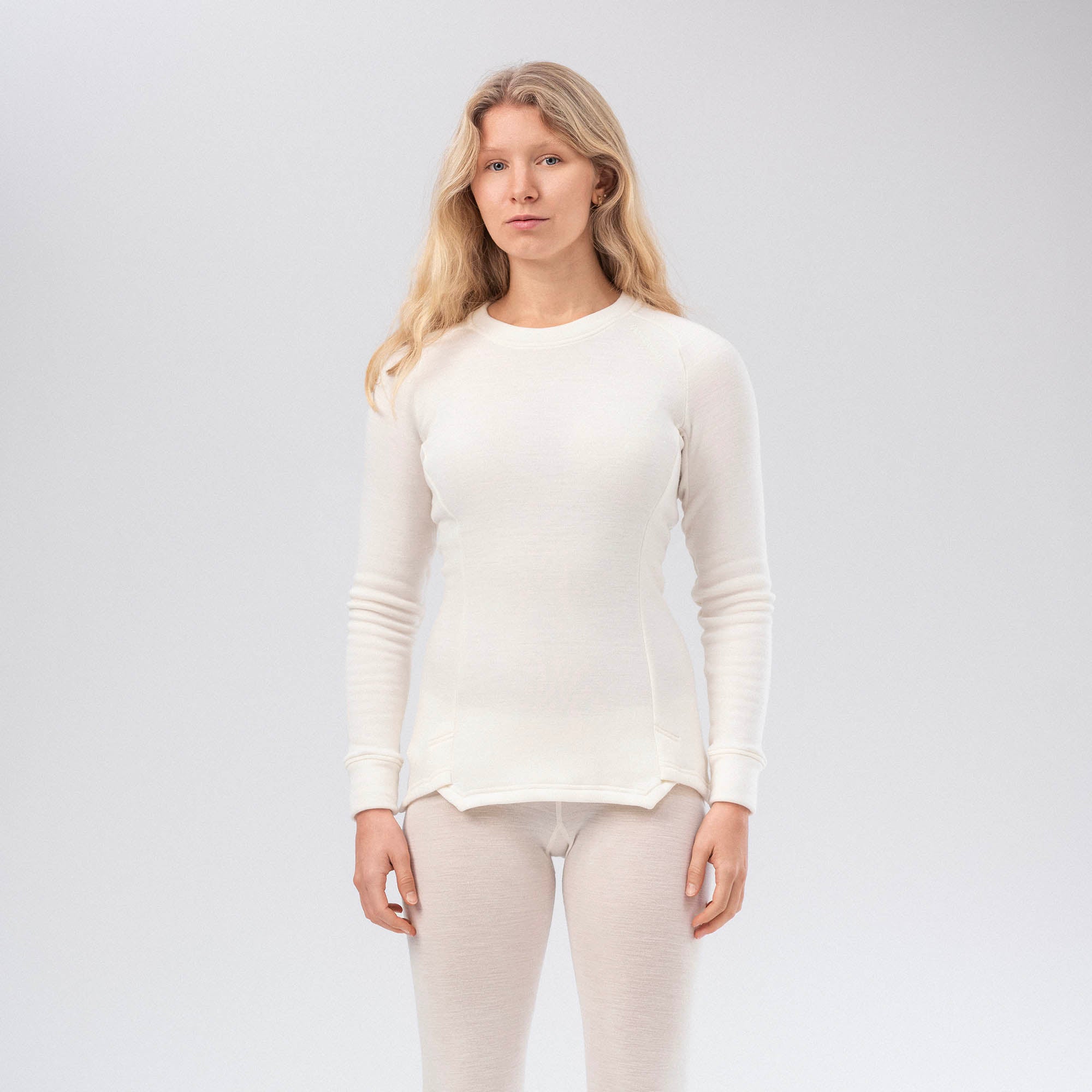 A woman with long blonde hair faces forward, wearing the Jersey Merino – AirWool 260 Women top and matching leggings in Marshmallow, set against a plain light background that emphasizes comfort and warmth.