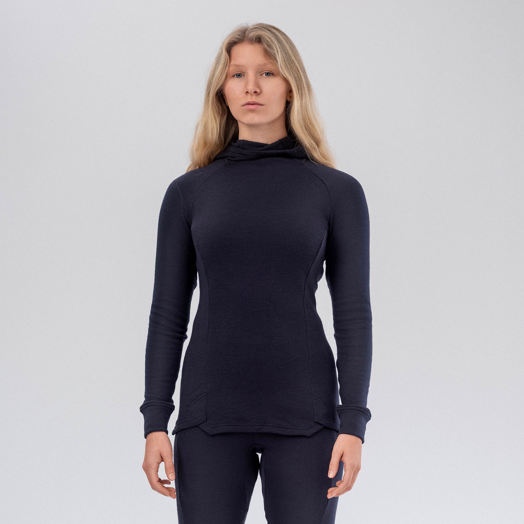 A woman with long blonde hair wears the Navy Hooded Jersey Merino – AirWool 260 Women and matching pants, standing against a plain light gray background.