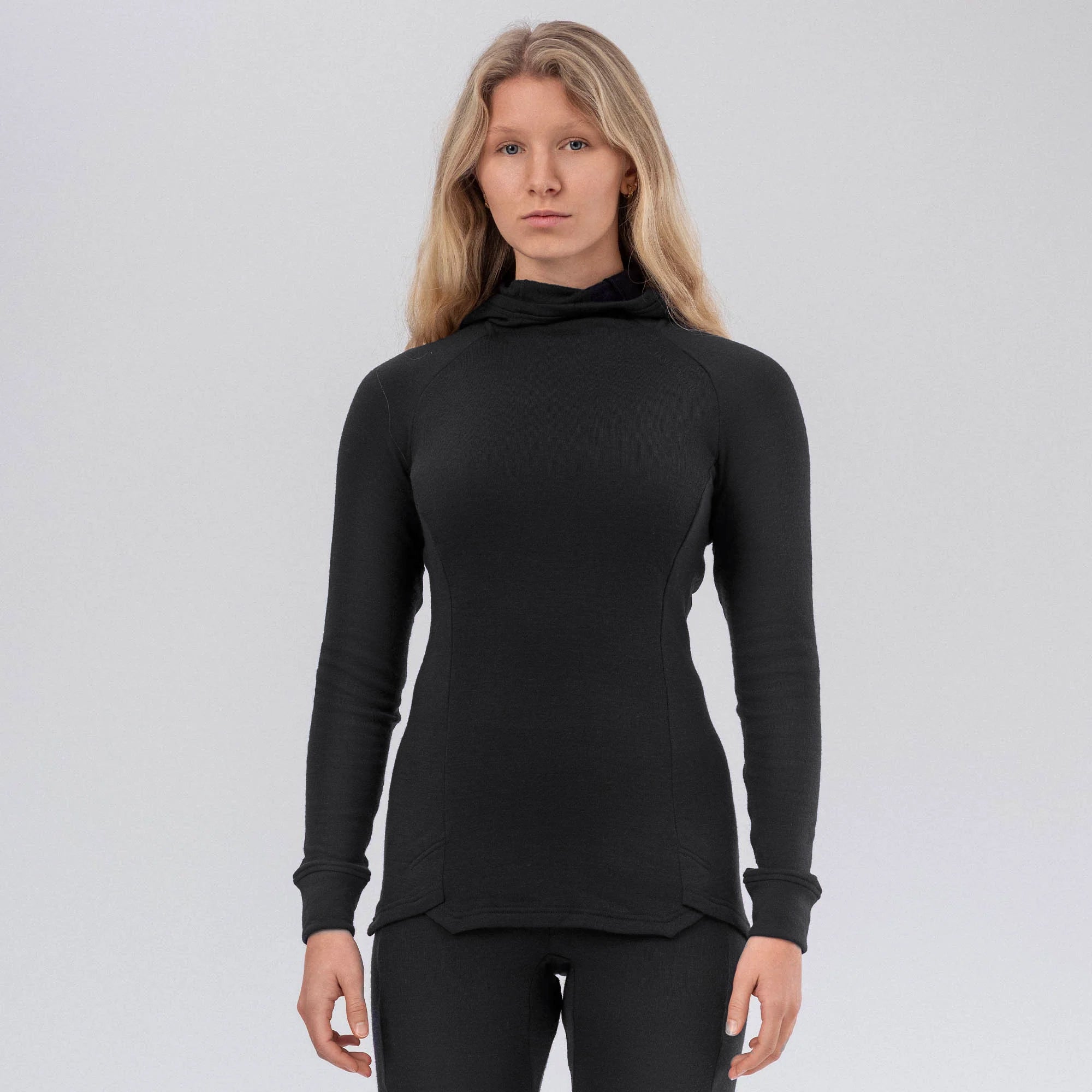 A woman with long blond hair wears the black Hooded Jersey Merino – AirWool 260 Women, standing against a plain light gray background.