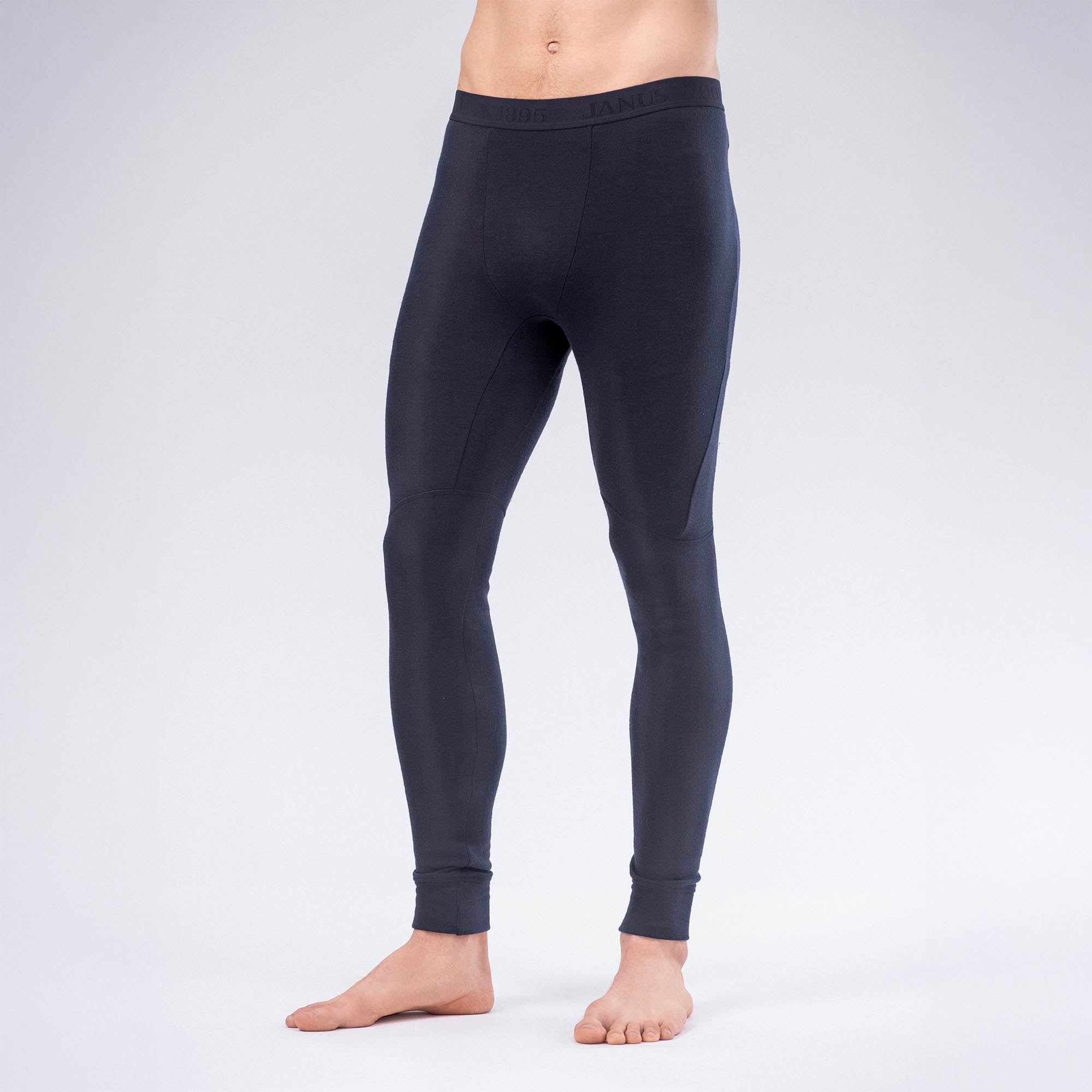 A person stands barefoot against a plain light background, wearing Leggings Merino – AirWool 260 Men. The dark, form-fitting merino wool leggings are full-length, covering from waist to ankles. The person's head is cropped out of the image.