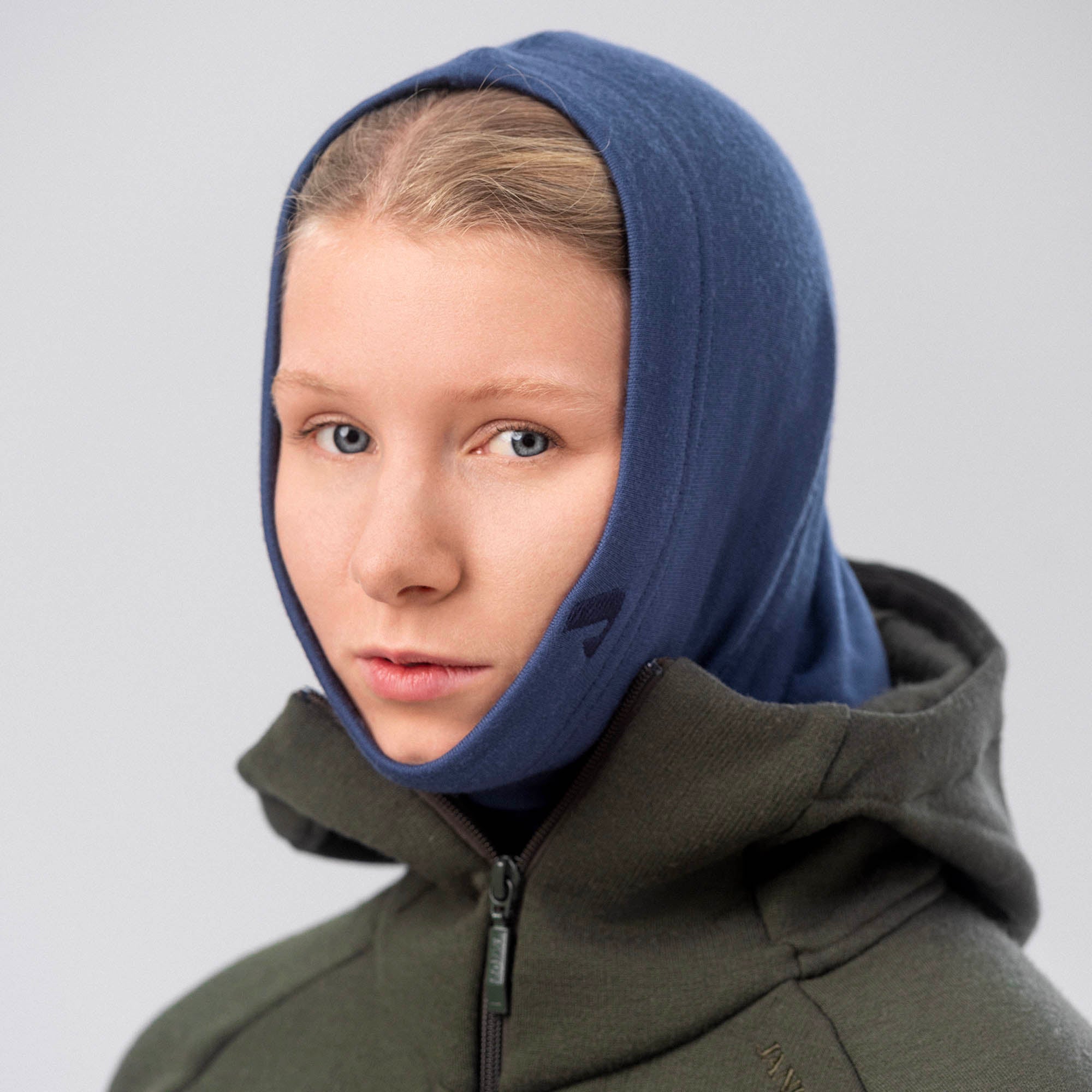 A person with light skin and blonde hair wears the Neck Warmer Merino – AirWool 260 in blue under a green zip-up jacket, facing slightly left with a neutral expression against a plain gray background.