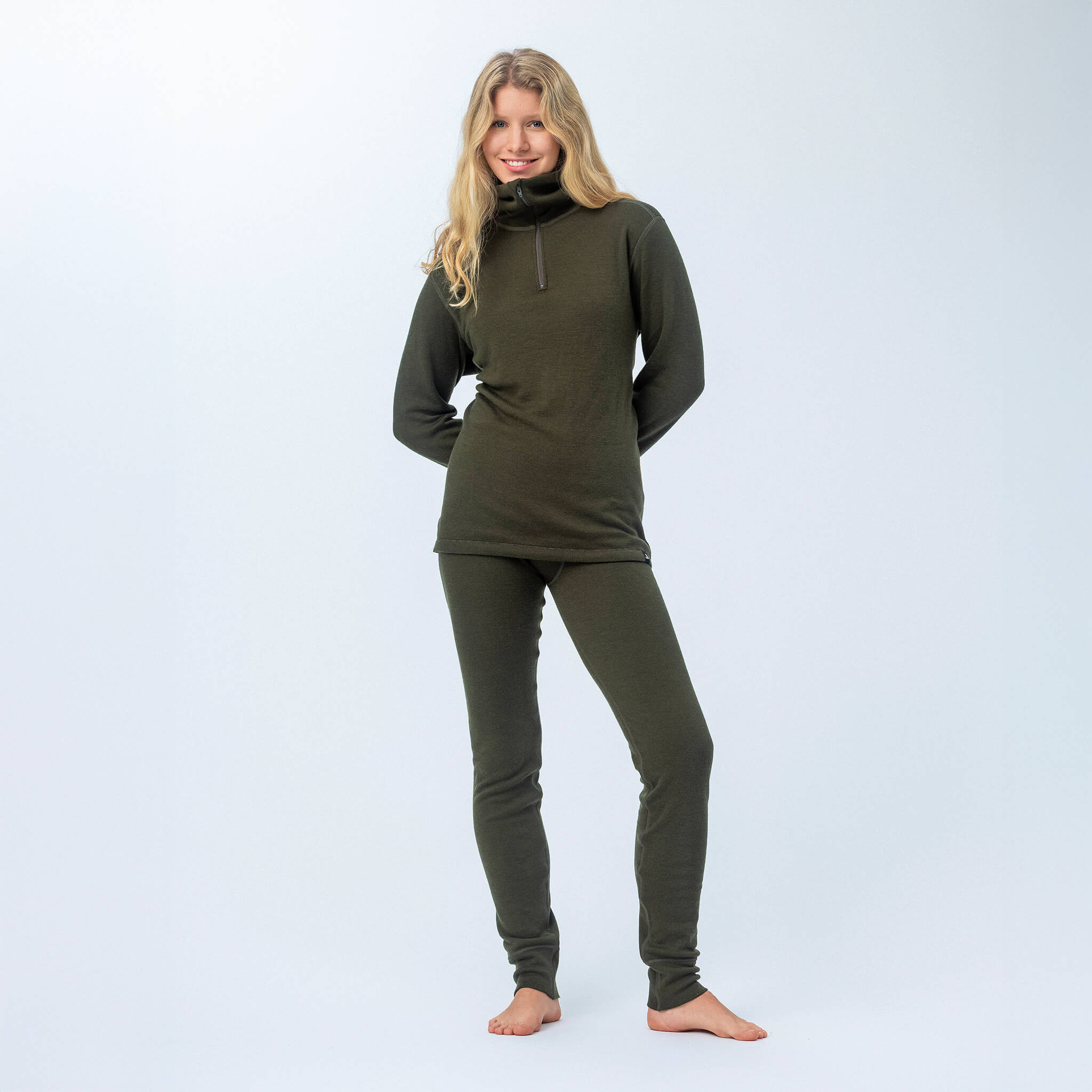 A person with long blonde hair stands barefoot against a plain light background, smiling with hands behind their back, wearing the Norse Merino 210 Unisex Polo with zip wool terry in dark green and fitted pants.