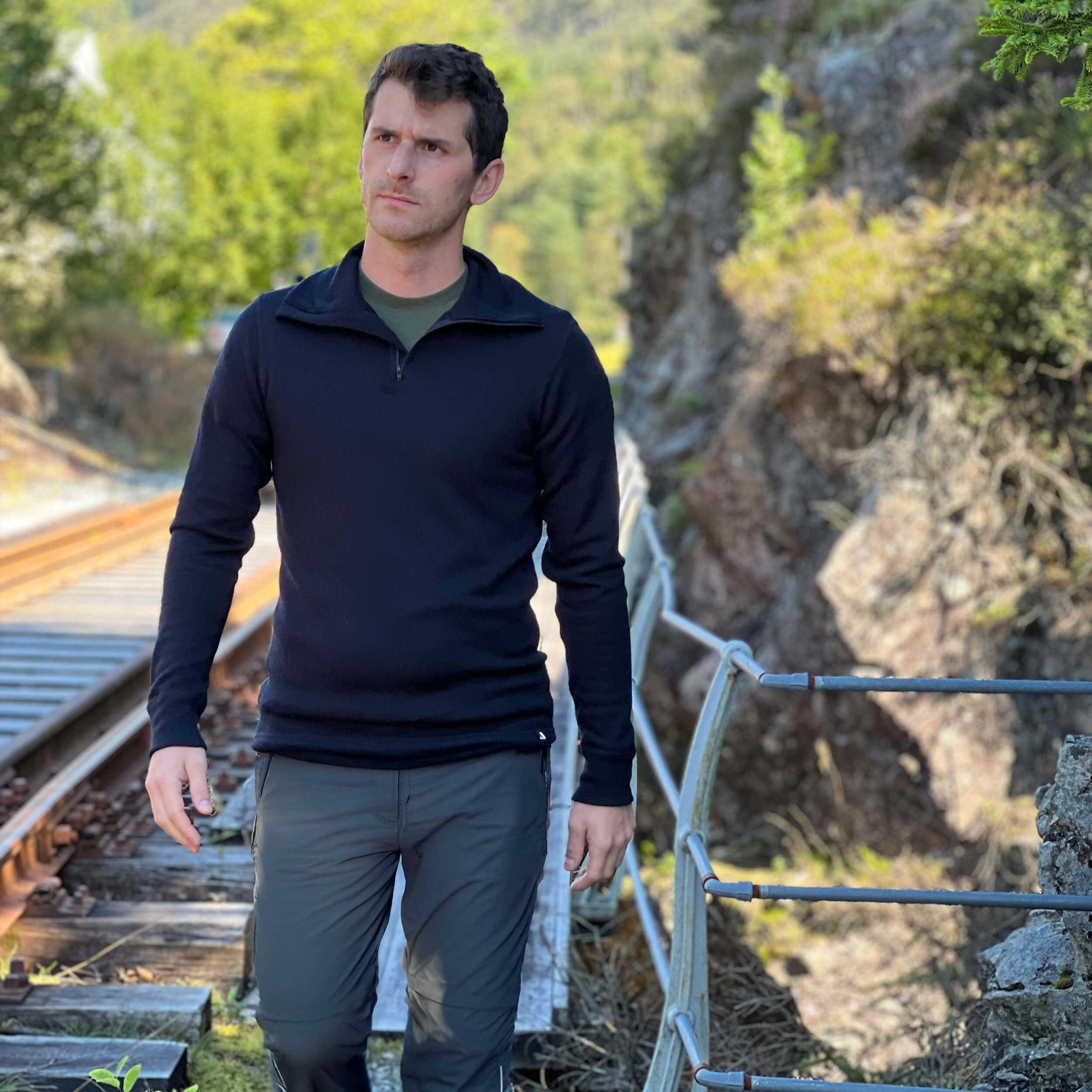 Man in Janus Norse Merino Polo in black, standing on a trail with greenery in the background