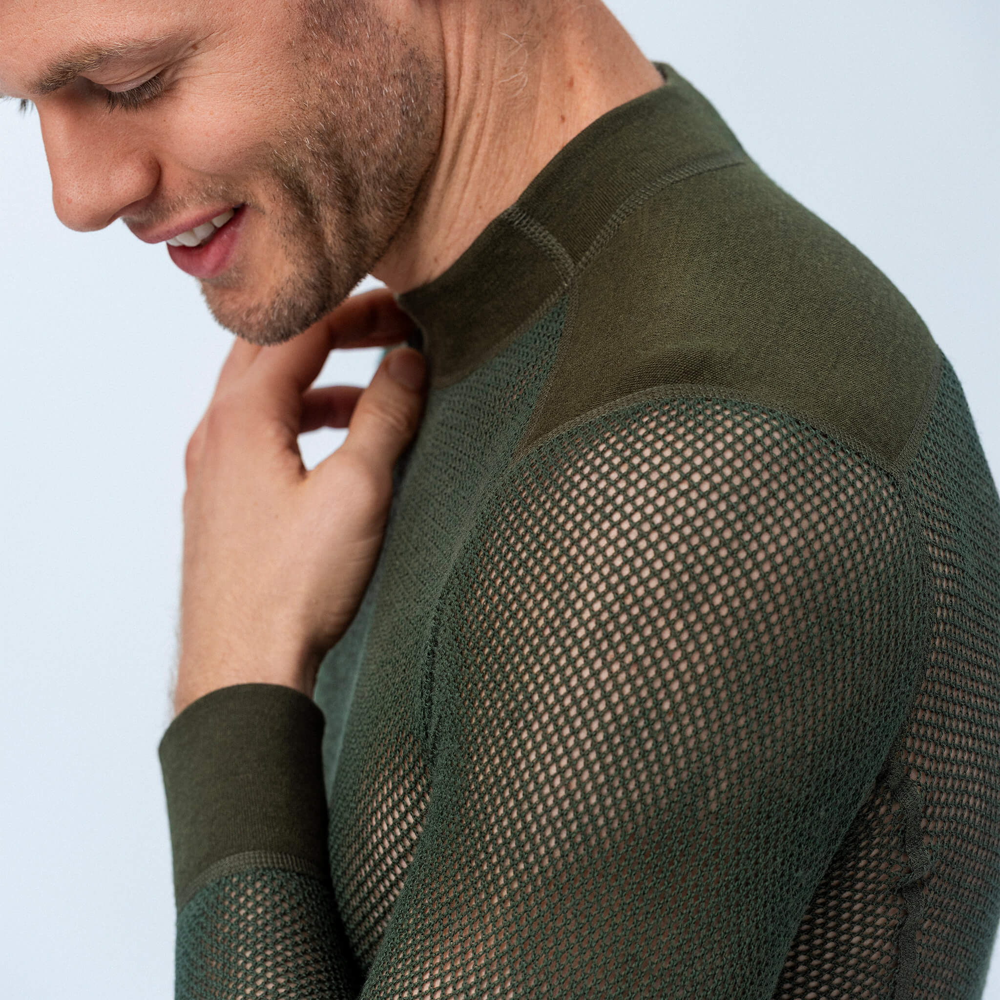 A smiling man, seen from the side against a light background, wears the Norse Merino 165 Unisex jersey wool mesh in olive green with long sleeves and reinforced elbows, touching his neck and looking down.