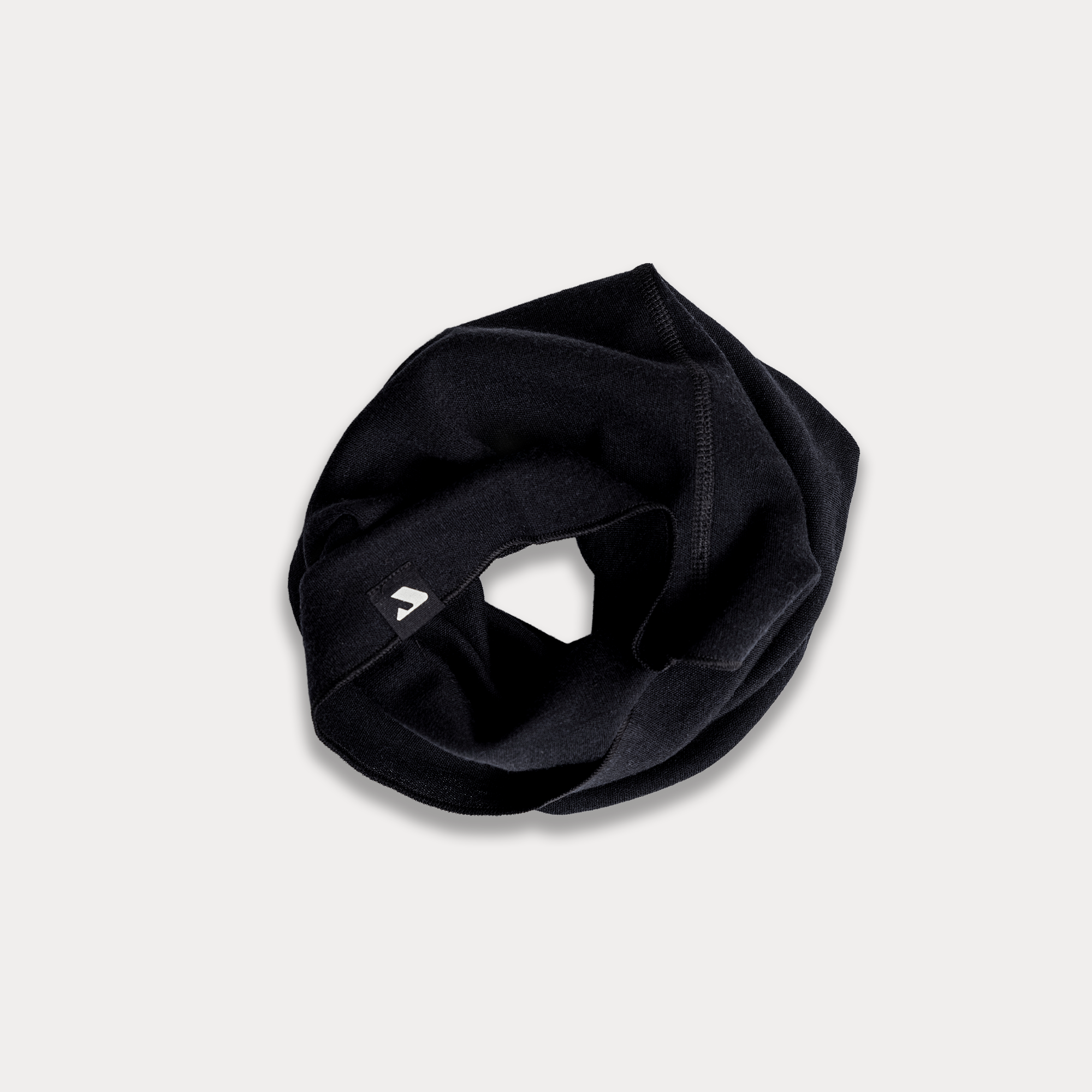 The Headover - Norse Merino 220, a black merino wool insulating neck gaiter or scarf, is placed in a loose circle on a light background, with a small white logo visible on the fabric's edge.