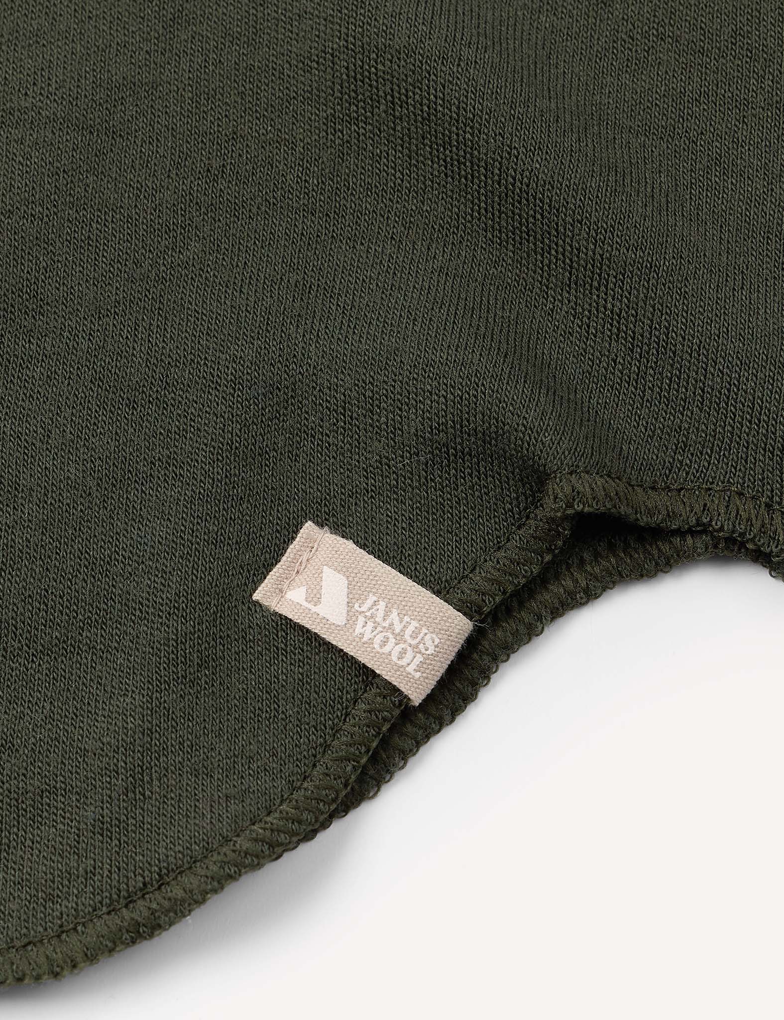 Close-up of the dark green knit fabric from the Balaclava – Merino Kids AirWool, featuring a small beige tag sewn into the seam. The fabric edge is slightly curved on a white background.