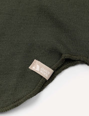 Close-up of the dark green knit fabric from the Balaclava – Merino Kids AirWool, featuring a small beige tag sewn into the seam. The fabric edge is slightly curved on a white background.