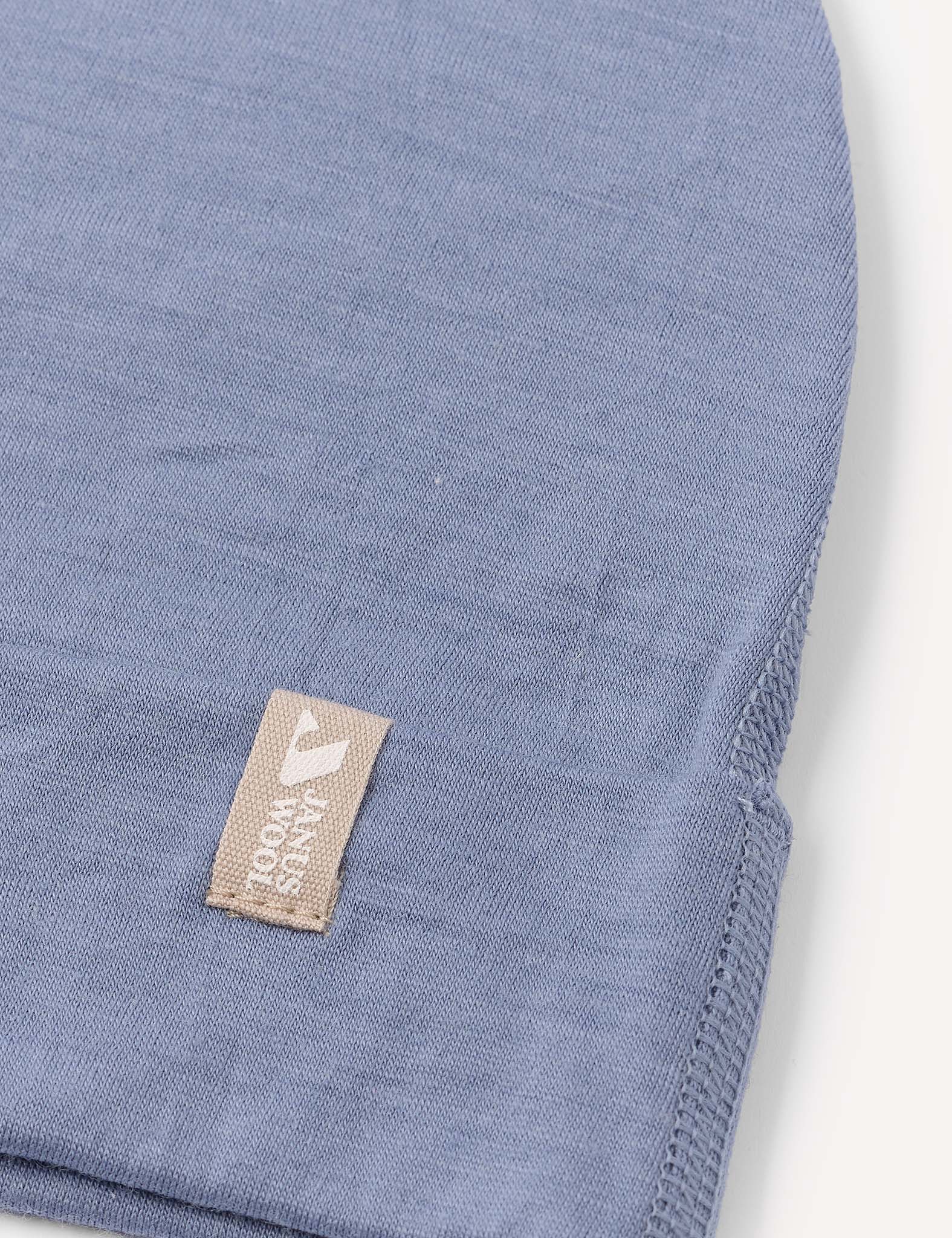 A close-up of the Beanie – Merino Kids Light in light blue, featuring a beige tag with white text and a logo. The hat’s soft merino wool material is evident, with visible stitching along the edge.