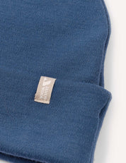 A close-up of the Beanie – Merino Kids AirWool in blue, featuring a beige tag with a white logo and text sewn onto the brim, set against a plain white background.