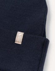 A close-up of the Beanie – Merino Kids AirWool in navy blue shows its soft, breathable merino wool fabric and a small beige tag with white text sewn onto the folded cuff.