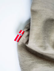 A close-up of breathable beige fabric with a small red, white, and blue Norwegian flag tag sewn into the seam, set on a white background. Ideal for the T-shirt Regular – Classic Merino 160 Men, known for temperature regulation.