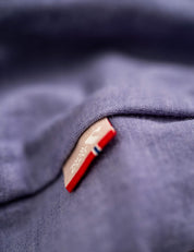 Close-up of a tan tag sewn onto soft blue Classic Merino 160 Men T-shirt fabric, featuring a small red and blue stripe on the tag’s edge. The breathable merino wool texture is clearly visible.