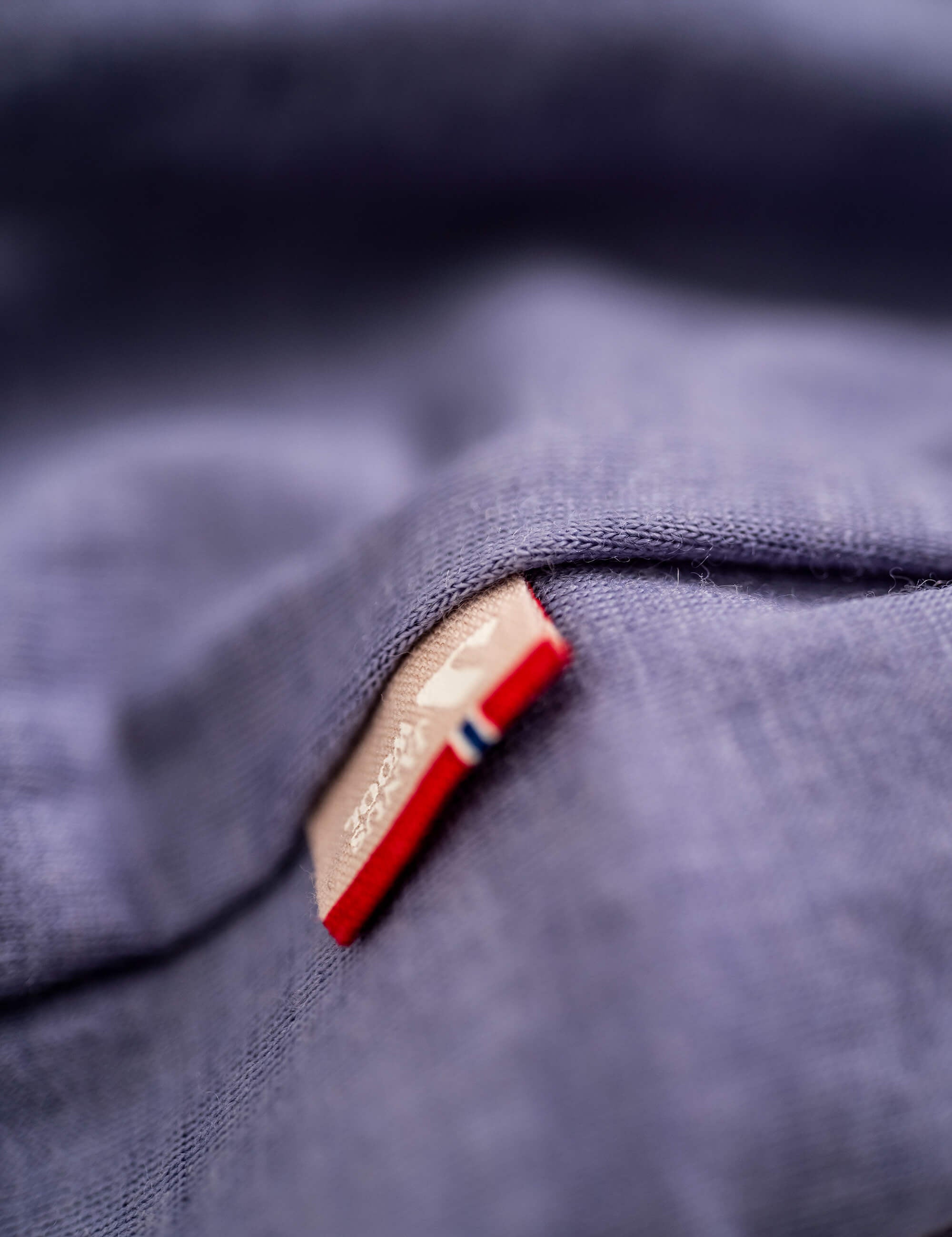 Close-up of a tan tag sewn onto soft blue Classic Merino 160 Men T-shirt fabric, featuring a small red and blue stripe on the tag’s edge. The breathable merino wool texture is clearly visible.