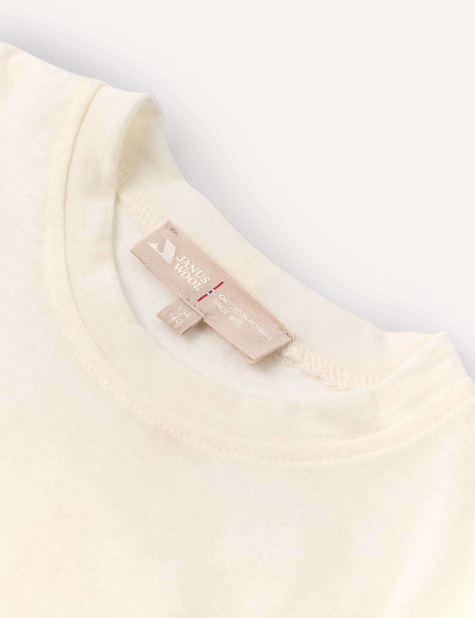 Close-up of the neckline of the Crewneck – Merino Kids Light shirt in a cream tone, crafted from soft merino wool jersey. A beige fabric tag featuring text and symbols is sewn into the inside collar. The background is light and uncluttered.