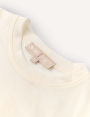 Close-up of the neckline of the Crewneck – Merino Kids Light shirt in a cream tone, crafted from soft merino wool jersey. A beige fabric tag featuring text and symbols is sewn into the inside collar. The background is light and uncluttered.