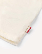 Close-up of the hem of the Crewneck – Merino Kids Light, a cream-colored t-shirt made from soft wool jersey, featuring a small beige and red fabric tag sewn into the seam.