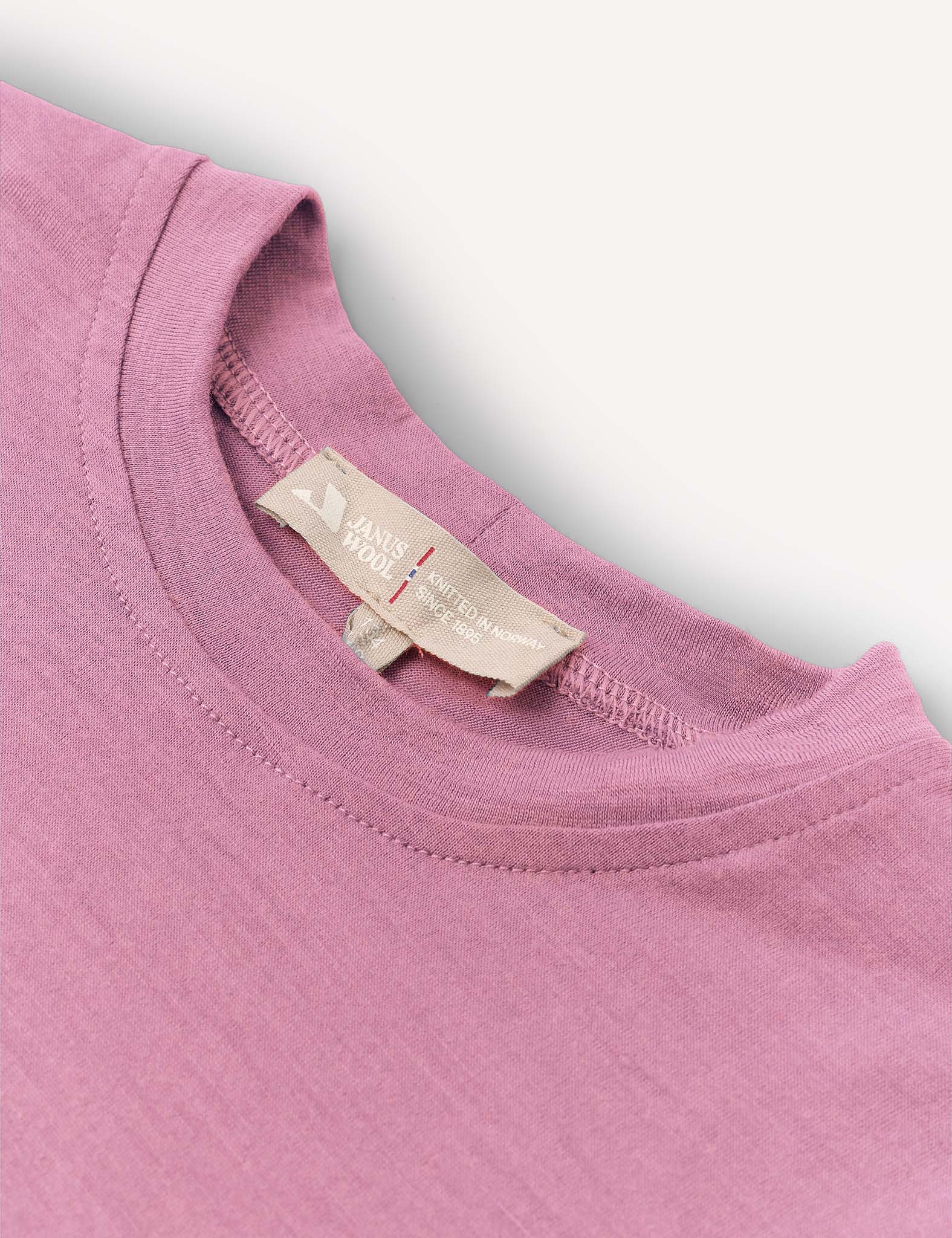 A close-up of the Crewneck – Merino Kids Light, a soft, pink crew neck t-shirt, focusing on the collar and tag featuring text and a small heart icon. The shirt is laid flat against a white background.