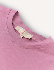 A close-up of the Crewneck – Merino Kids Light, a soft, pink crew neck t-shirt, focusing on the collar and tag featuring text and a small heart icon. The shirt is laid flat against a white background.