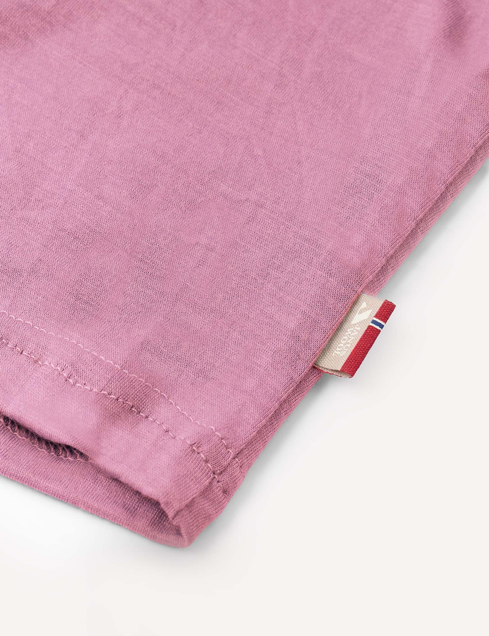 Close-up of the hem of the Crewneck – Merino Kids Light in pink soft wool jersey, featuring a small beige tag with a red, white, and blue stripe detail on a light background.