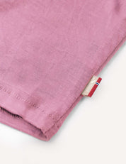 Close-up of the hem of the Crewneck – Merino Kids Light in pink soft wool jersey, featuring a small beige tag with a red, white, and blue stripe detail on a light background.
