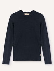 A navy “Crewneck – Merino Kids Light” long-sleeved shirt made from soft merino wool is laid flat on a light background. It features a small white and red tag on the lower left side near the hem.