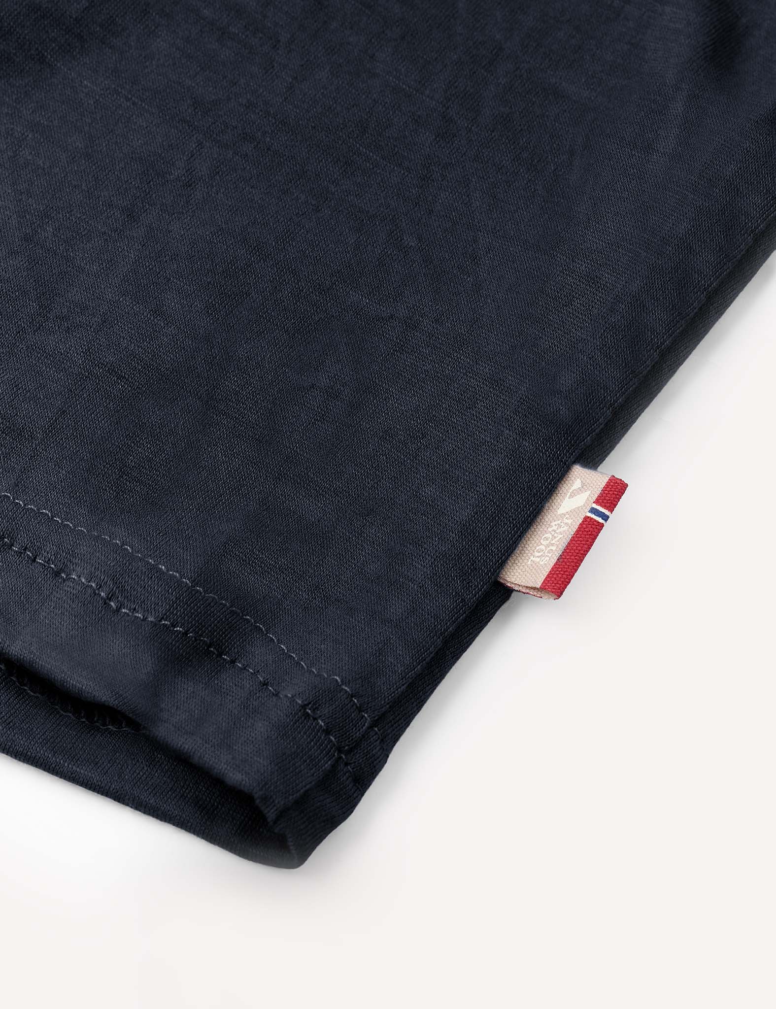 Close-up of the hem of the Crewneck – Merino Kids Light in dark navy merino wool, featuring a small tag with a red, white, and blue stripe and some text, set against a plain light background.
