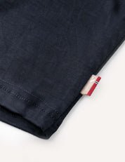 Close-up of the hem of the Crewneck – Merino Kids Light in dark navy merino wool, featuring a small tag with a red, white, and blue stripe and some text, set against a plain light background.