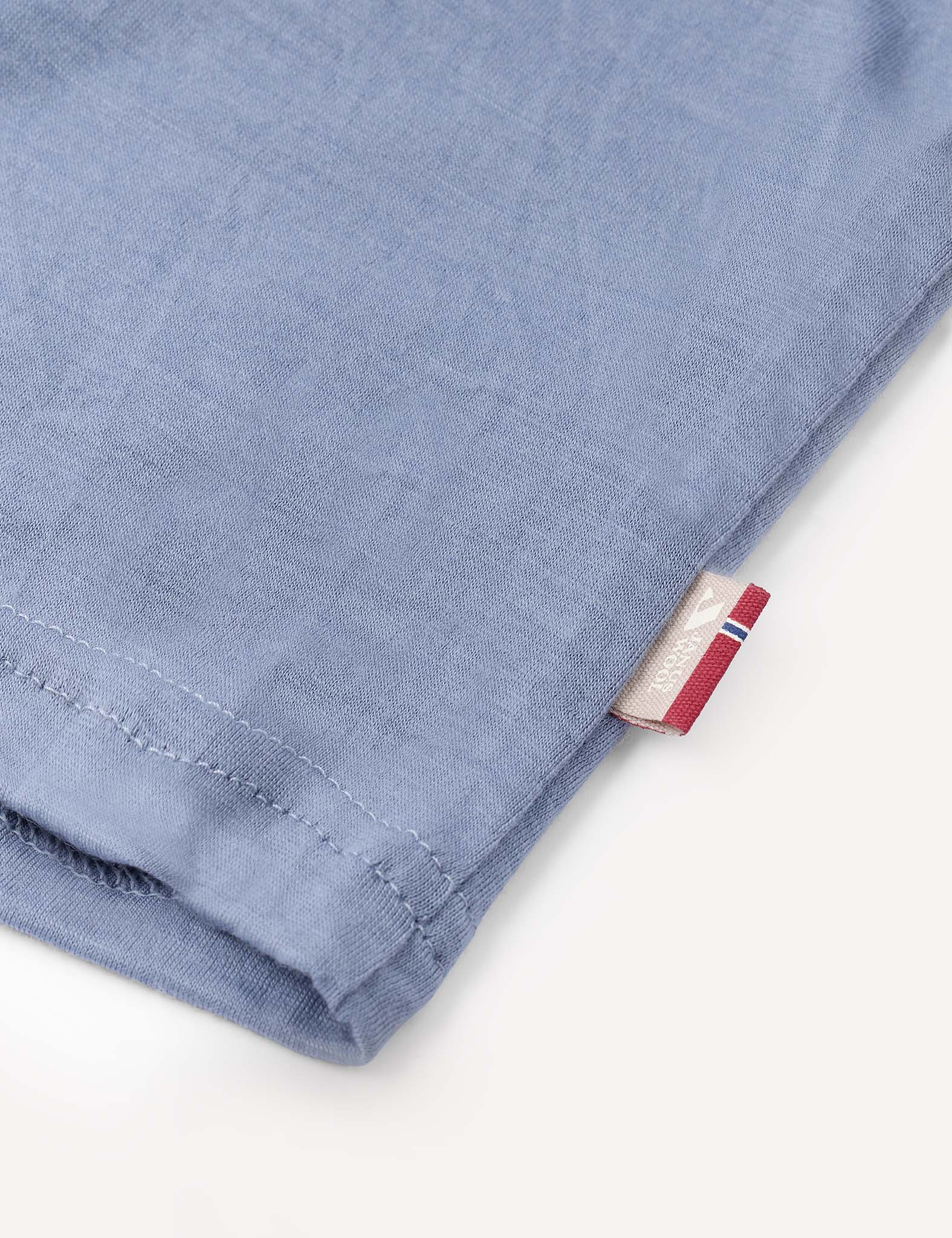 Close-up of the soft, light blue fabric of the Crewneck – Merino Kids Light, featuring a small white tag with red and blue detailing sewn into the seam. The background is a plain light surface.