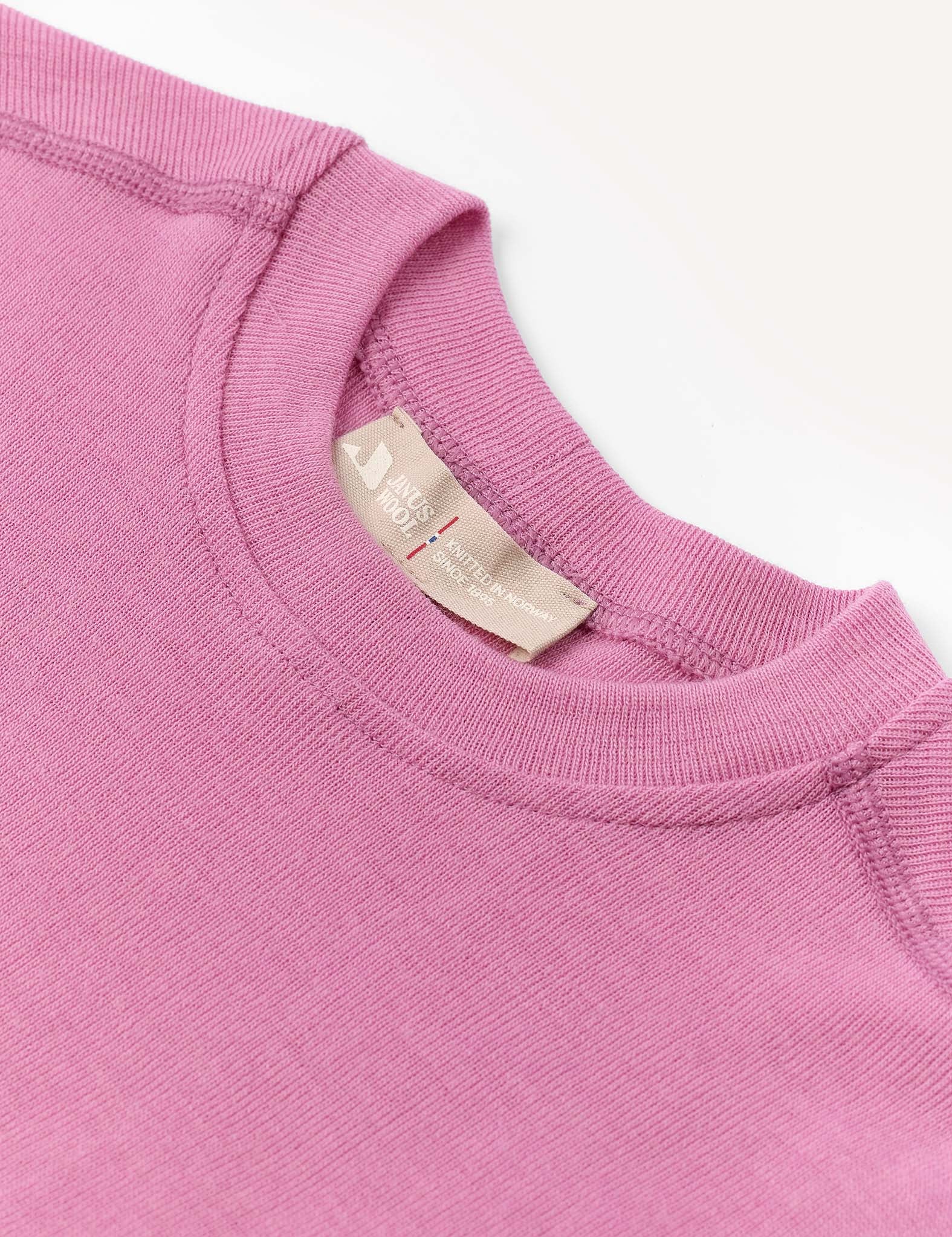 A close-up of the Crewneck – Merino Kids Classic Rib in pink, crafted from ribbed wool jersey, featuring visible stitching and a beige fabric tag at the neckline, displayed against a white background.