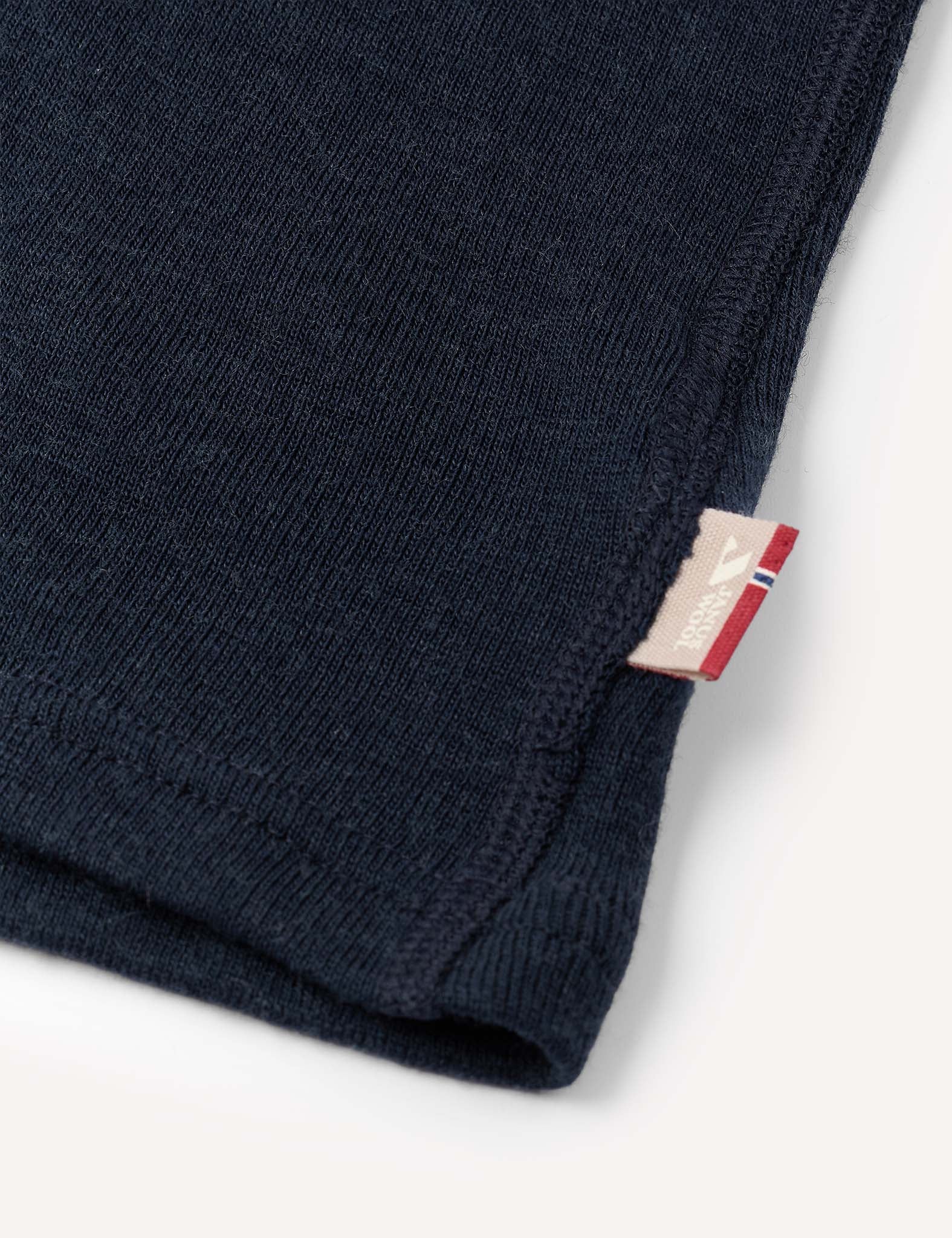 Close-up of the corner of the Crewneck – Merino Kids Classic Rib in dark blue ribbed wool jersey, featuring a small beige and red tag sewn into the seam, set against a plain off-white background.