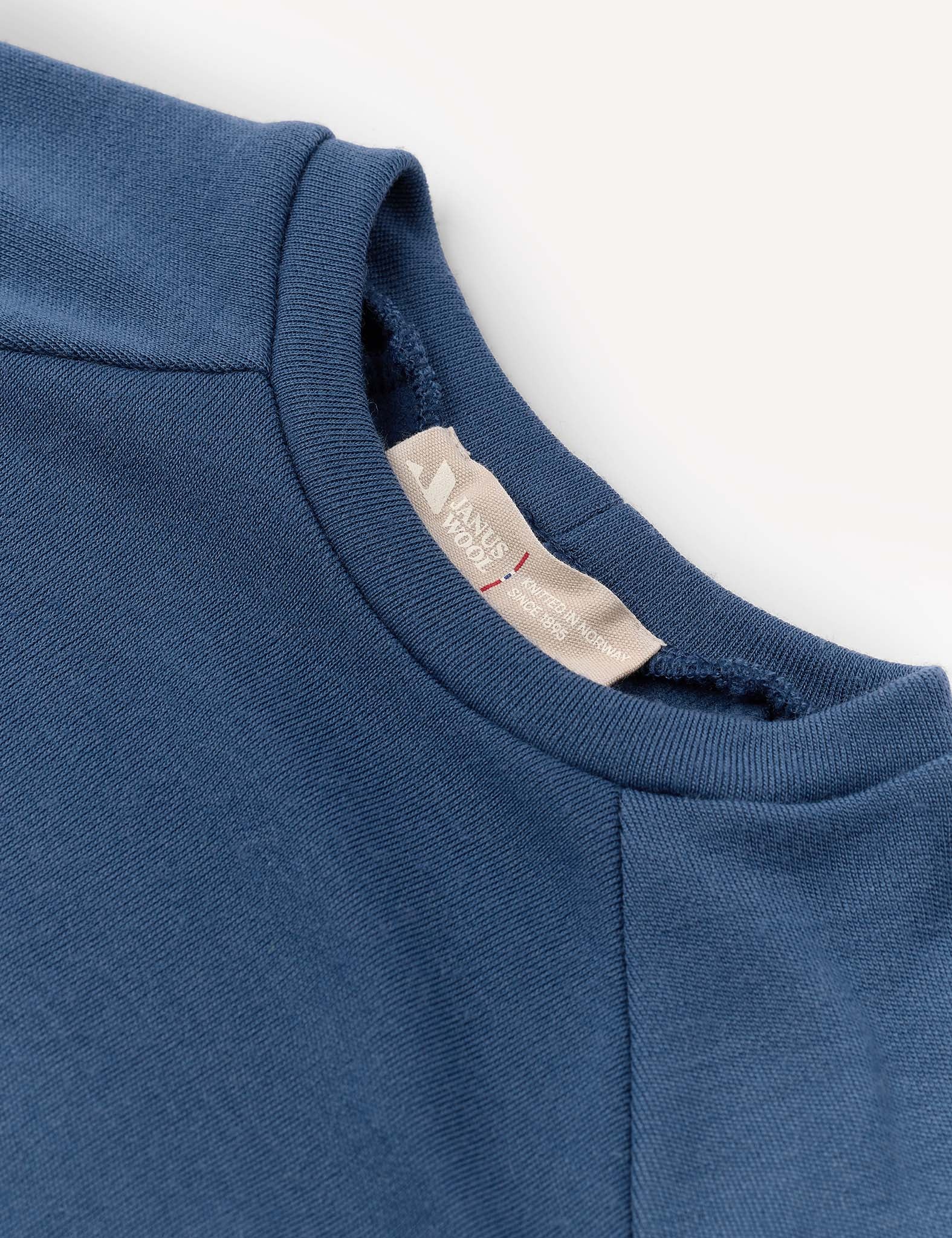 A close-up of the blue Crewneck – Merino Kids AirWool sweatshirt highlights the collar area and inner tag, which reads HANDY WARDROBE. The fabric looks soft with a subtle texture.