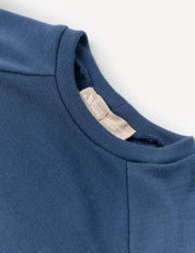 A close-up of the blue Crewneck – Merino Kids AirWool sweatshirt highlights the collar area and inner tag, which reads HANDY WARDROBE. The fabric looks soft with a subtle texture.