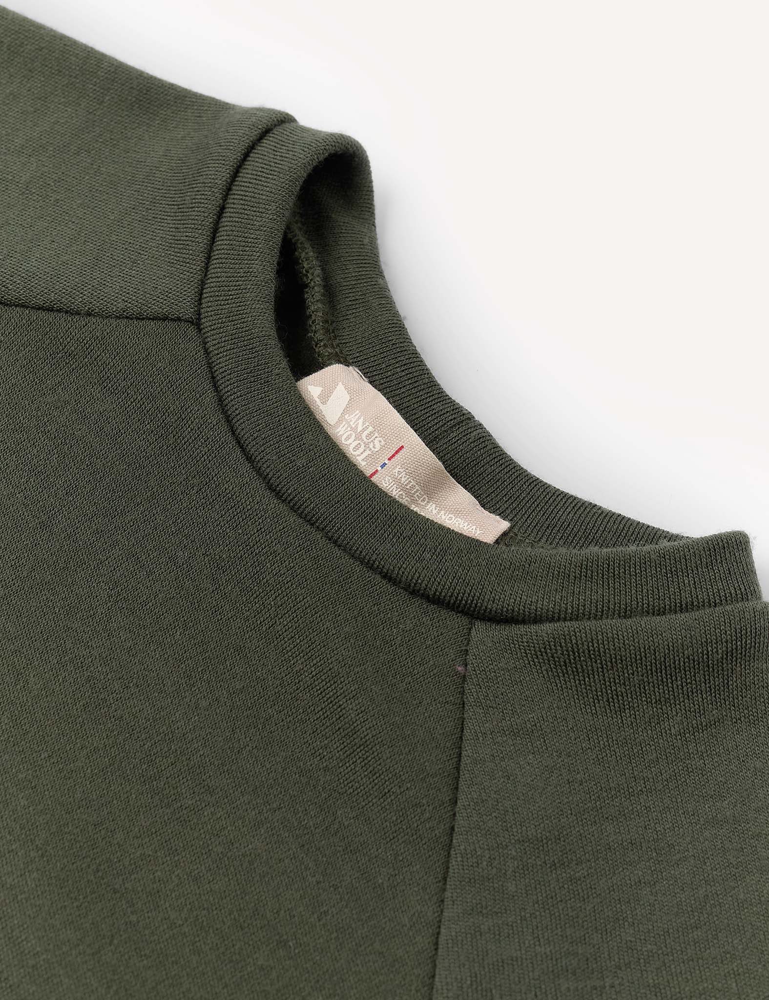 Close-up of the Crewneck – Merino Kids AirWool sweatshirt in dark green, showcasing its soft merino wool AirWool 260 fabric, detailed crewneck collar, and a beige fabric tag with text. The background is clean and light-colored.