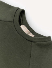 Close-up of the Crewneck – Merino Kids AirWool sweatshirt in dark green, showcasing its soft merino wool AirWool 260 fabric, detailed crewneck collar, and a beige fabric tag with text. The background is clean and light-colored.