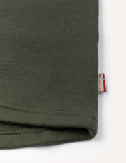 Close-up of the hem of the Crewneck – Merino Kids AirWool in olive green, featuring a small beige and red tag sewn into the seam, shown against a white background.