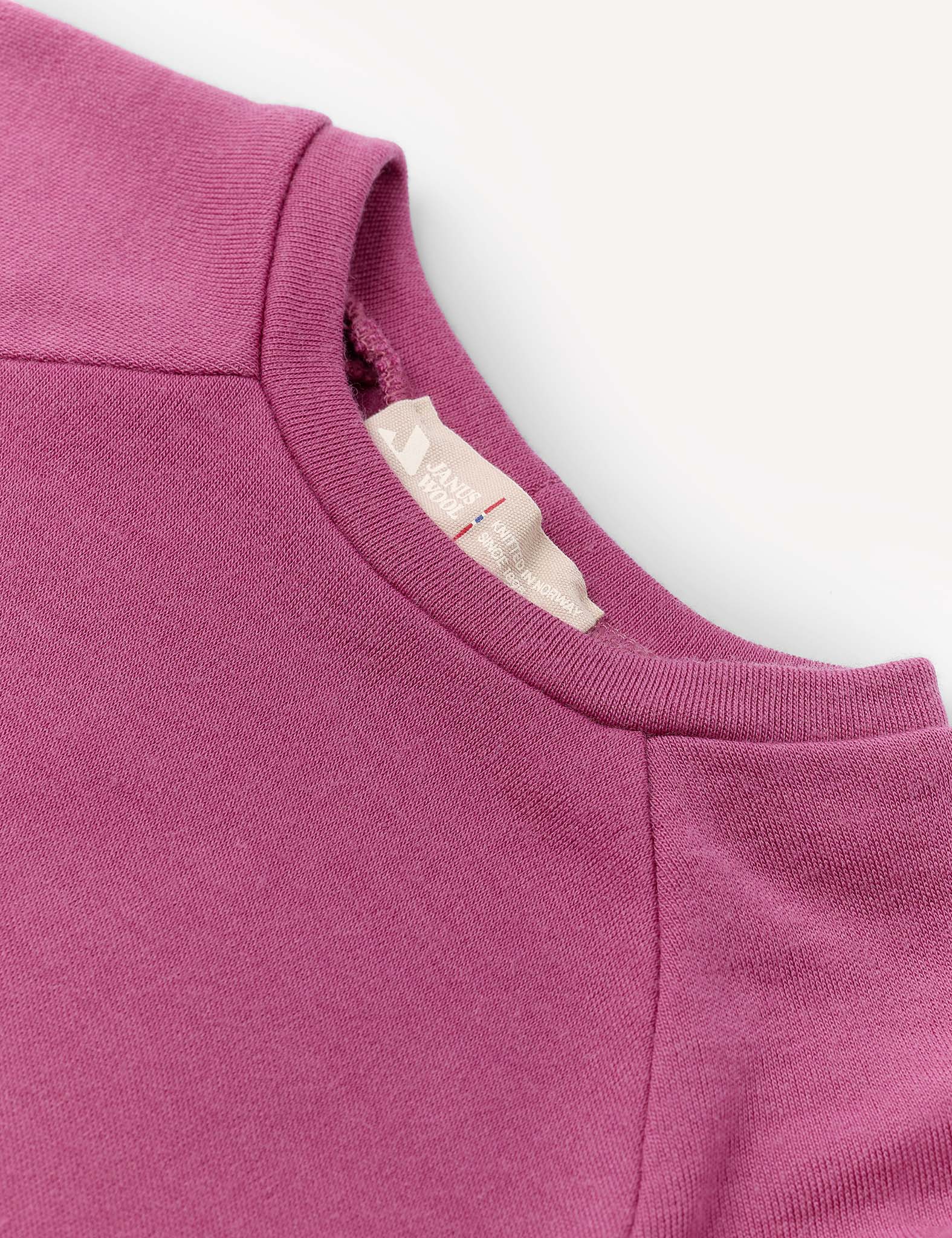 A close-up of the Crewneck – Merino Kids AirWool jersey sweatshirt in magenta showcases the collar, shoulder seams, and a beige inner neckline tag with text, all set against a light background. Crafted from AirWool 260 merino wool, this piece emphasizes premium comfort and warmth.