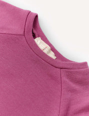 A close-up of the Crewneck – Merino Kids AirWool jersey sweatshirt in magenta showcases the collar, shoulder seams, and a beige inner neckline tag with text, all set against a light background. Crafted from AirWool 260 merino wool, this piece emphasizes premium comfort and warmth.