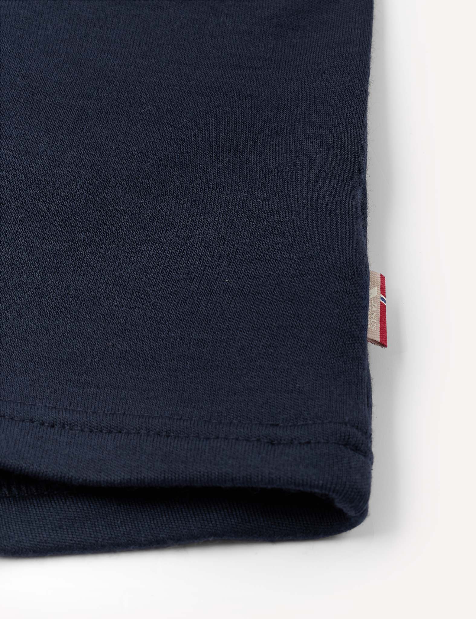 Close-up of the Crewneck – Merino Kids AirWool in navy blue merino wool, featuring a small red and white tag sewn into the seam, possibly showing a sleeve or pant leg, set against a plain white background.