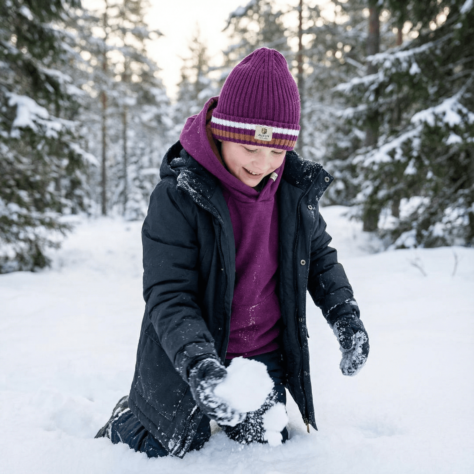 A child in a purple hat and Janus Wool's Polo with zip merino - Janus Go 250 Kids sweater kneels in snowy woods, smiling while making a snowball among the trees.