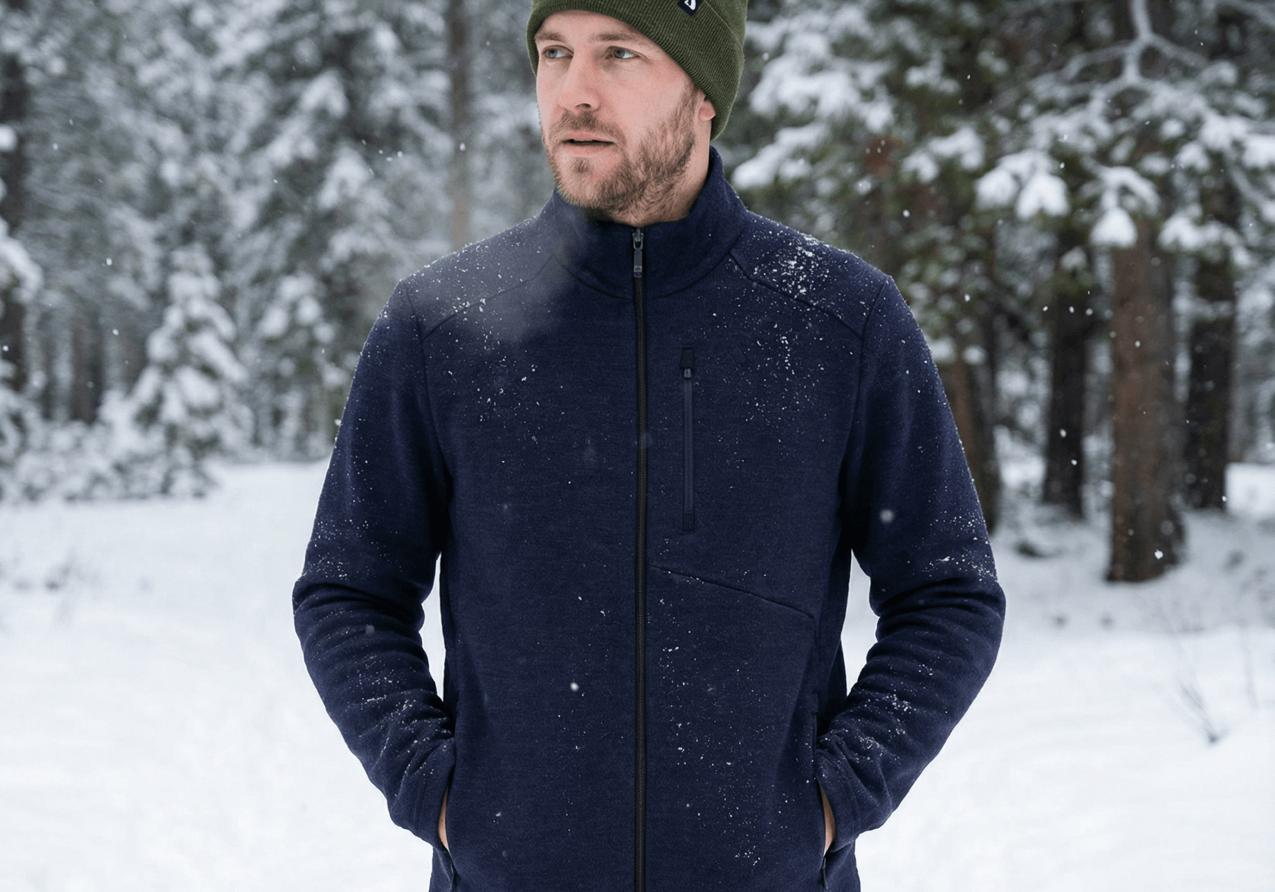 A man wearing a green merino wool beanie and navy jacket stands outdoors in a snowy forest, with trees and fresh snow in the background. He has his hands in his jacket pockets and looks slightly to the side.