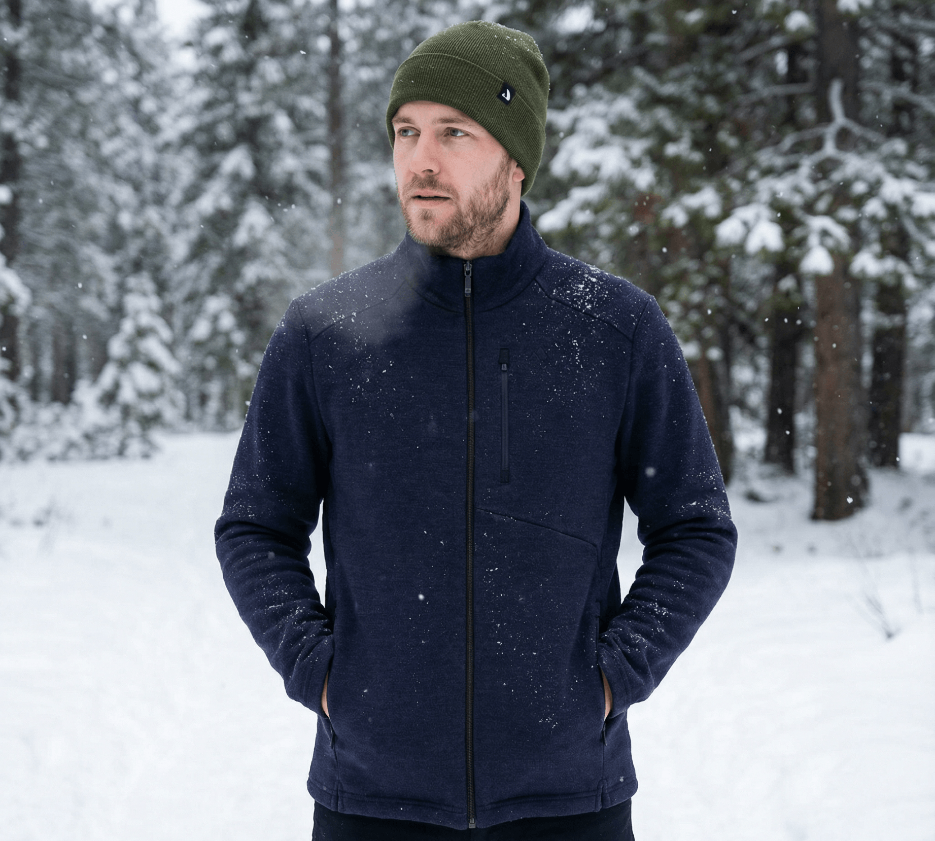 A man wearing a green merino wool beanie and navy jacket stands outdoors in a snowy forest, with trees and fresh snow in the background. He has his hands in his jacket pockets and looks slightly to the side.