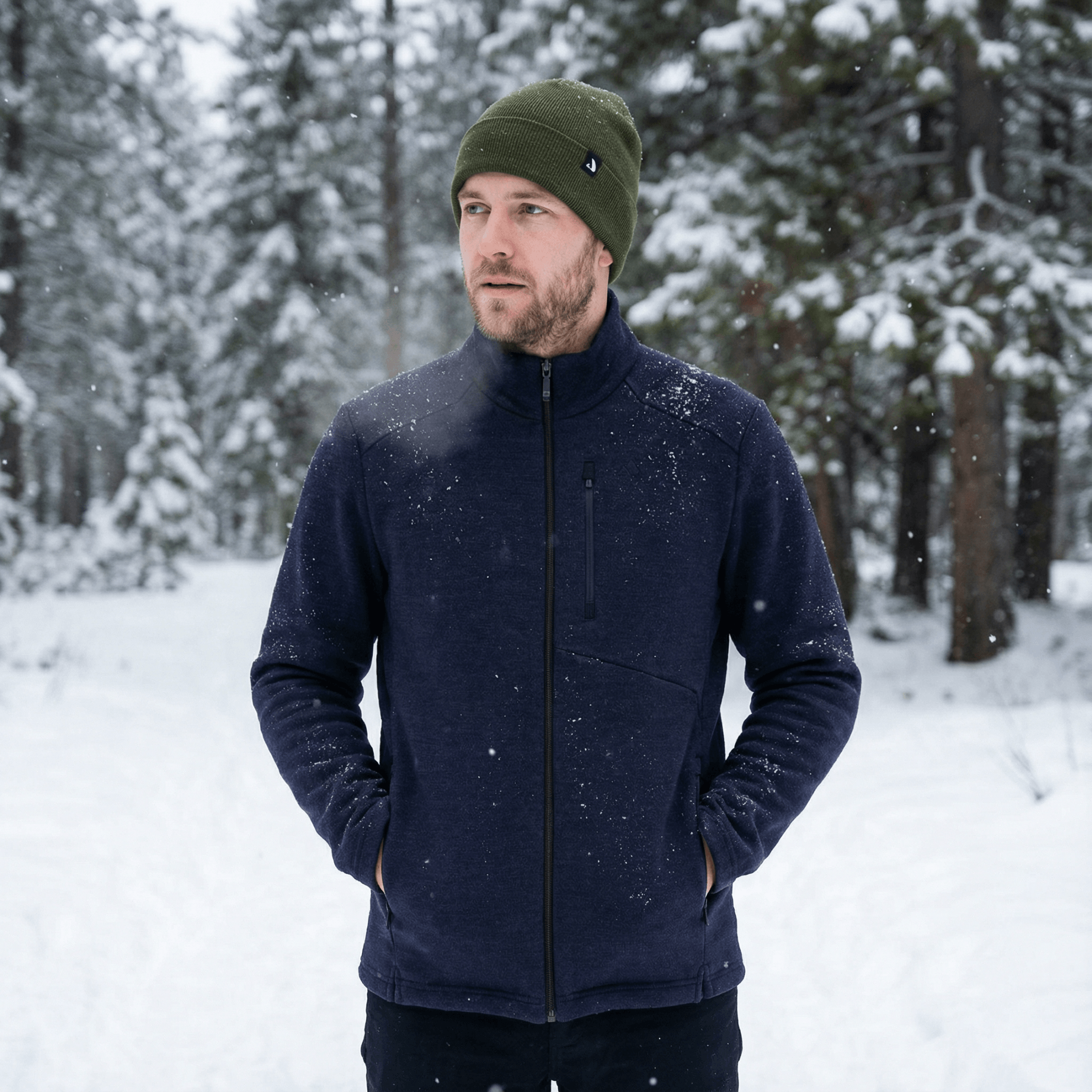 A man wearing a green merino wool beanie and navy jacket stands outdoors in a snowy forest, with trees and fresh snow in the background. He has his hands in his jacket pockets and looks slightly to the side.
