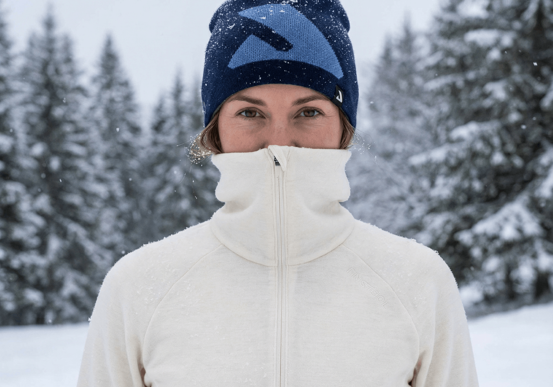 A person in a cream merino wool zip-up jacket and blue winter hat stands outside in falling snow, surrounded by snow-covered trees, with their jacket zipped up to their nose.