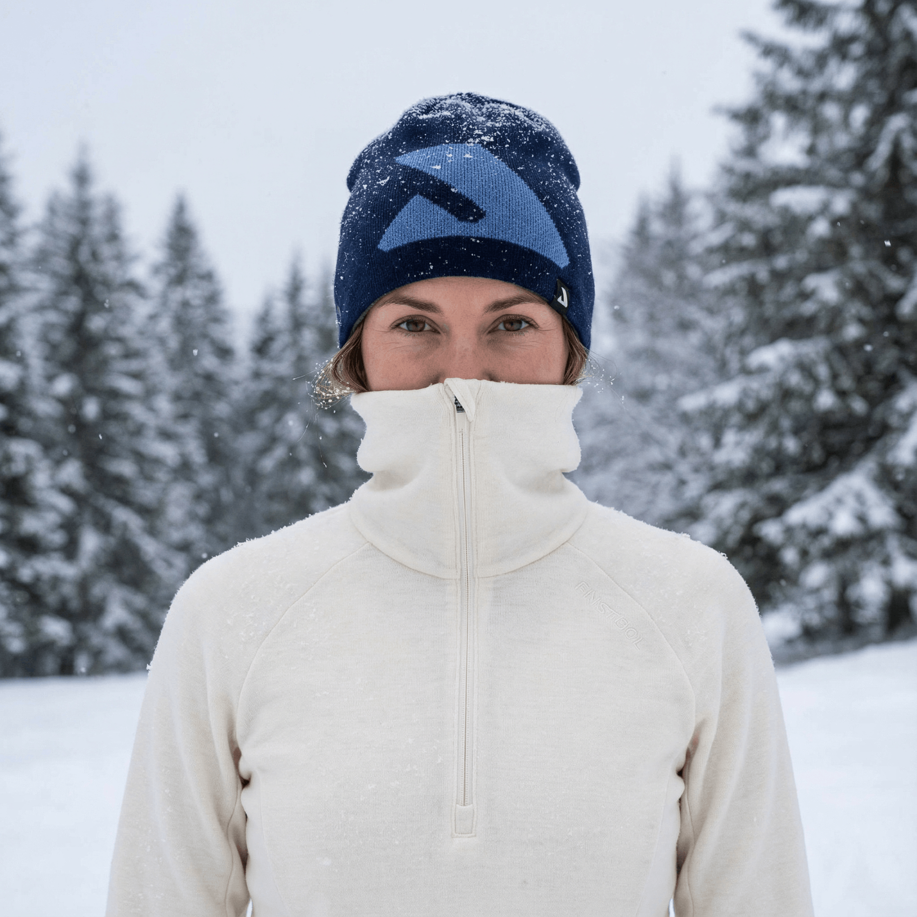 A person in a cream merino wool zip-up jacket and blue winter hat stands outside in falling snow, surrounded by snow-covered trees, with their jacket zipped up to their nose.