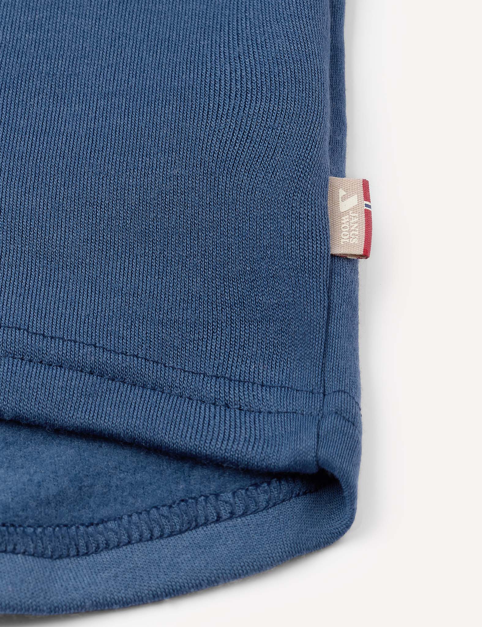 Close-up of the blue knit fabric of the Half Zip – Merino Kids AirWool, featuring a small beige tag with red, white, and blue stripes and text sewn into the edge seam.