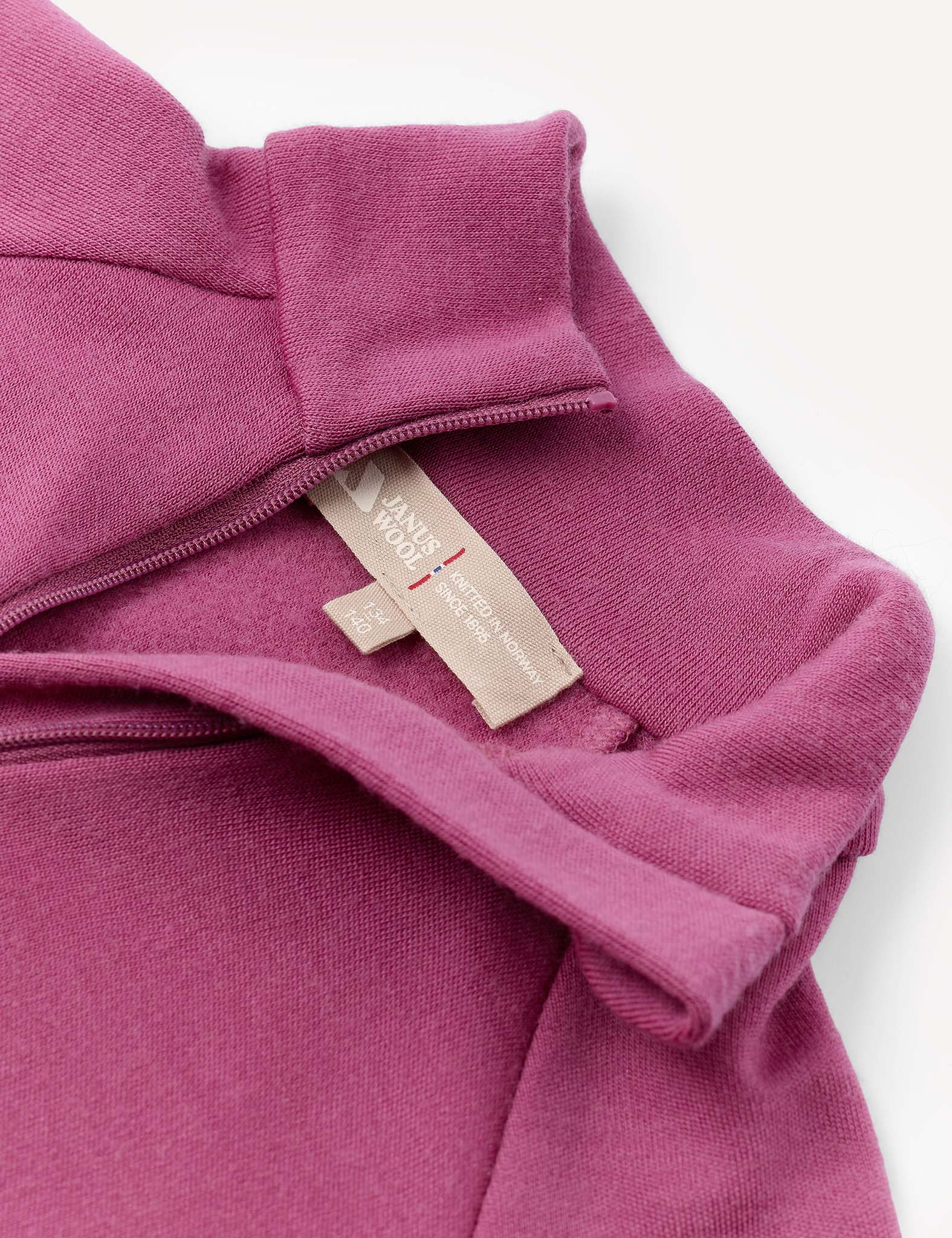 Close-up of a mauve Half Zip – Merino Kids AirWool garment featuring a brushed finish and half-zip collar, with two beige fabric labels visible: one marked 100% WOOL and the other displaying care and sizing information.