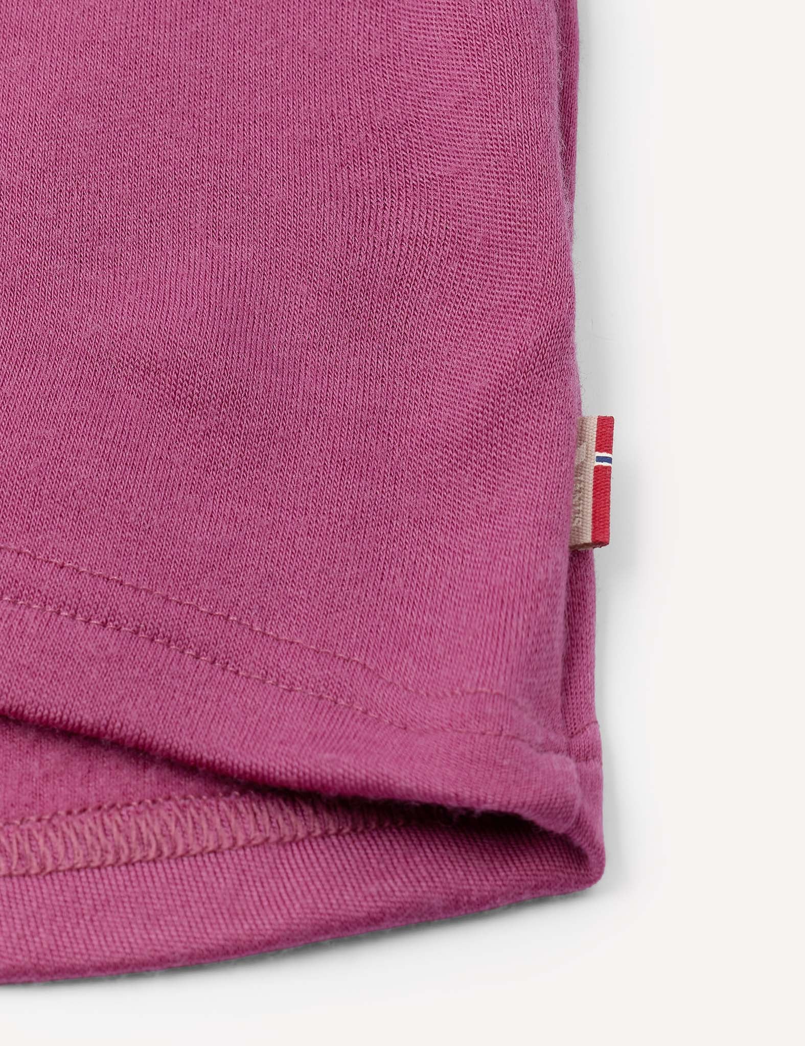 Close-up of the hem of the Half Zip – Merino Kids AirWool in pink, featuring a small fabric tag with red, white, and beige stripes sewn into the seam. The background is off-white.